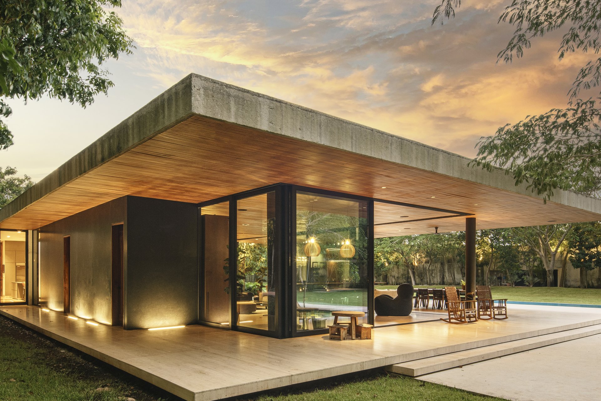 Casa Akúun, en Mérida, diseñada por Arkham Projects: la terraza cubierta de madera abierta al jardín y la alberca, con el interior iluminado al atardecer