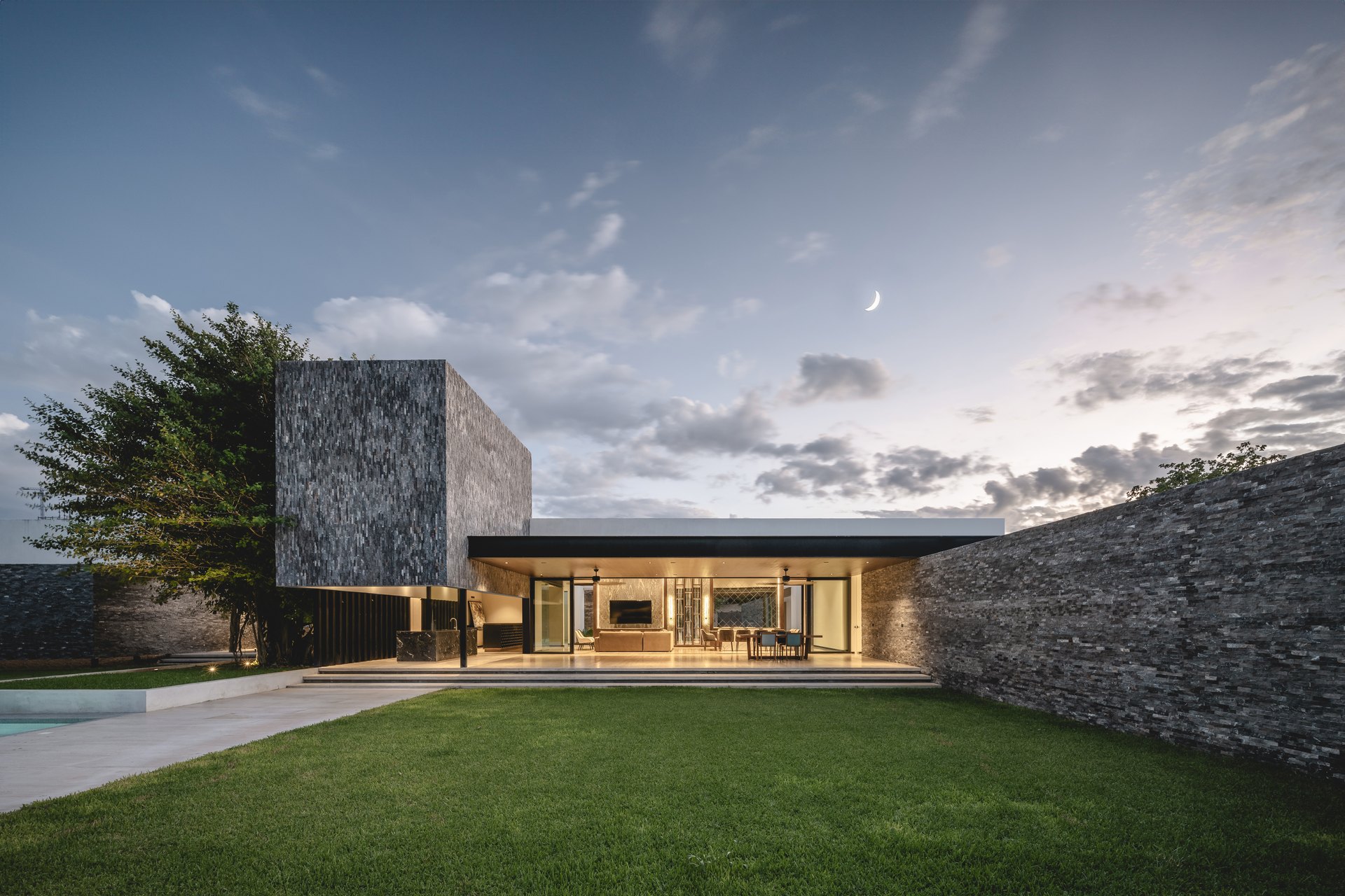 Casa Donna, en Mérida, diseñada por Arkham Projects: el jardín y la terraza abierta bajo el volumen de piedra, con el muro perimetral al atardecer