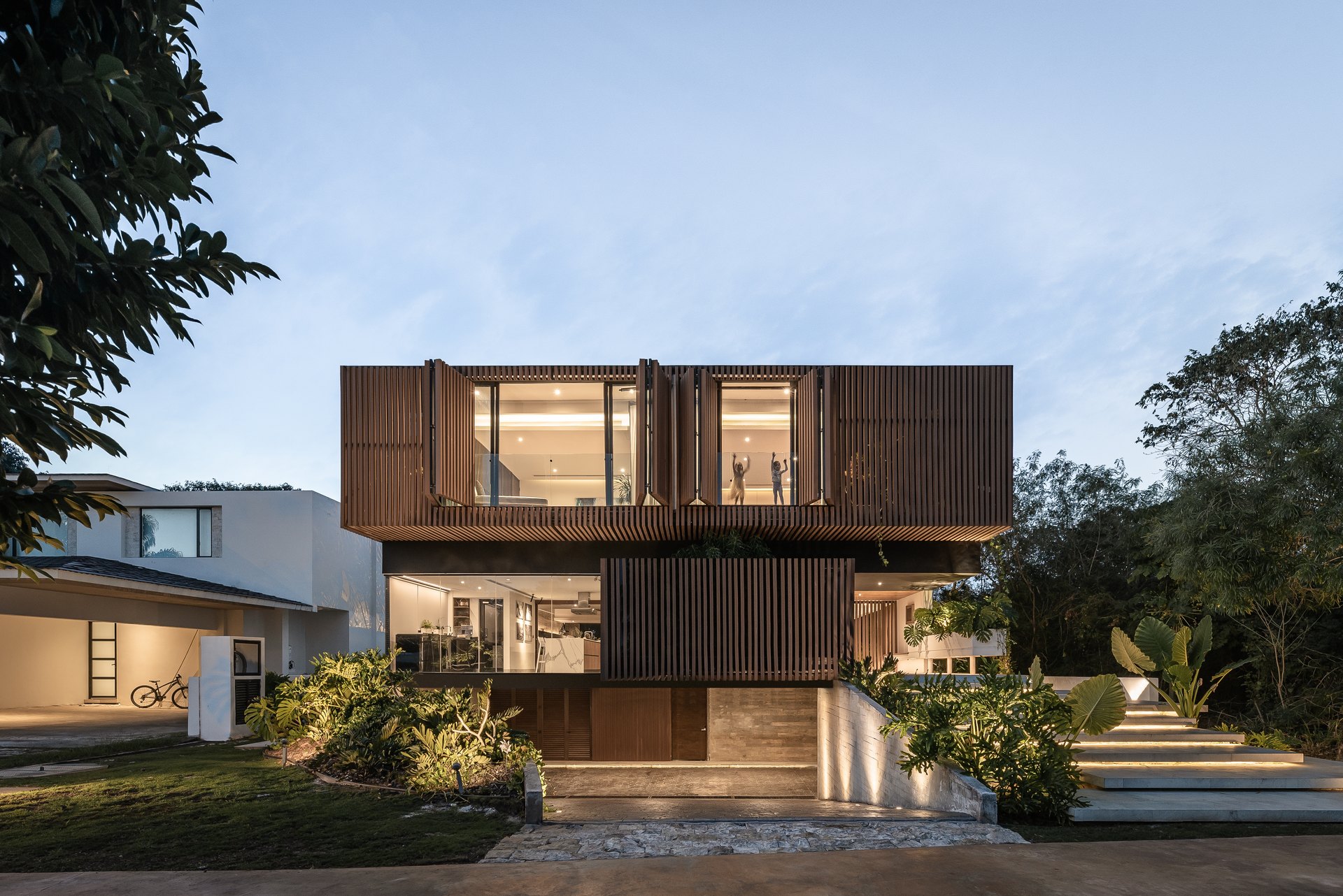 Casa Mulix, en Mérida, diseñada por Arkham Projects: la fachada con el volumen de celosía de madera flotando sobre el primer nivel, entre los árboles del terreno