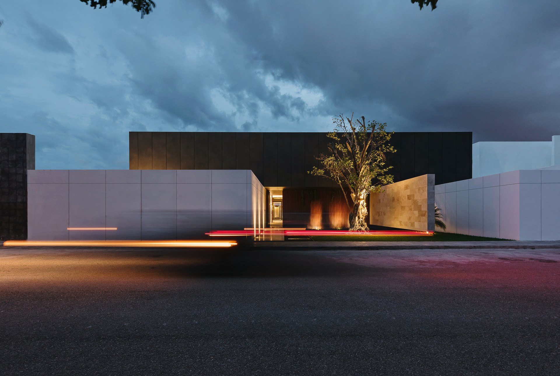 Casa Tmzn, en Mérida, diseñada por Arkham Projects: la fachada de acceso de volúmenes oscuros y claros con un árbol al centro, de noche